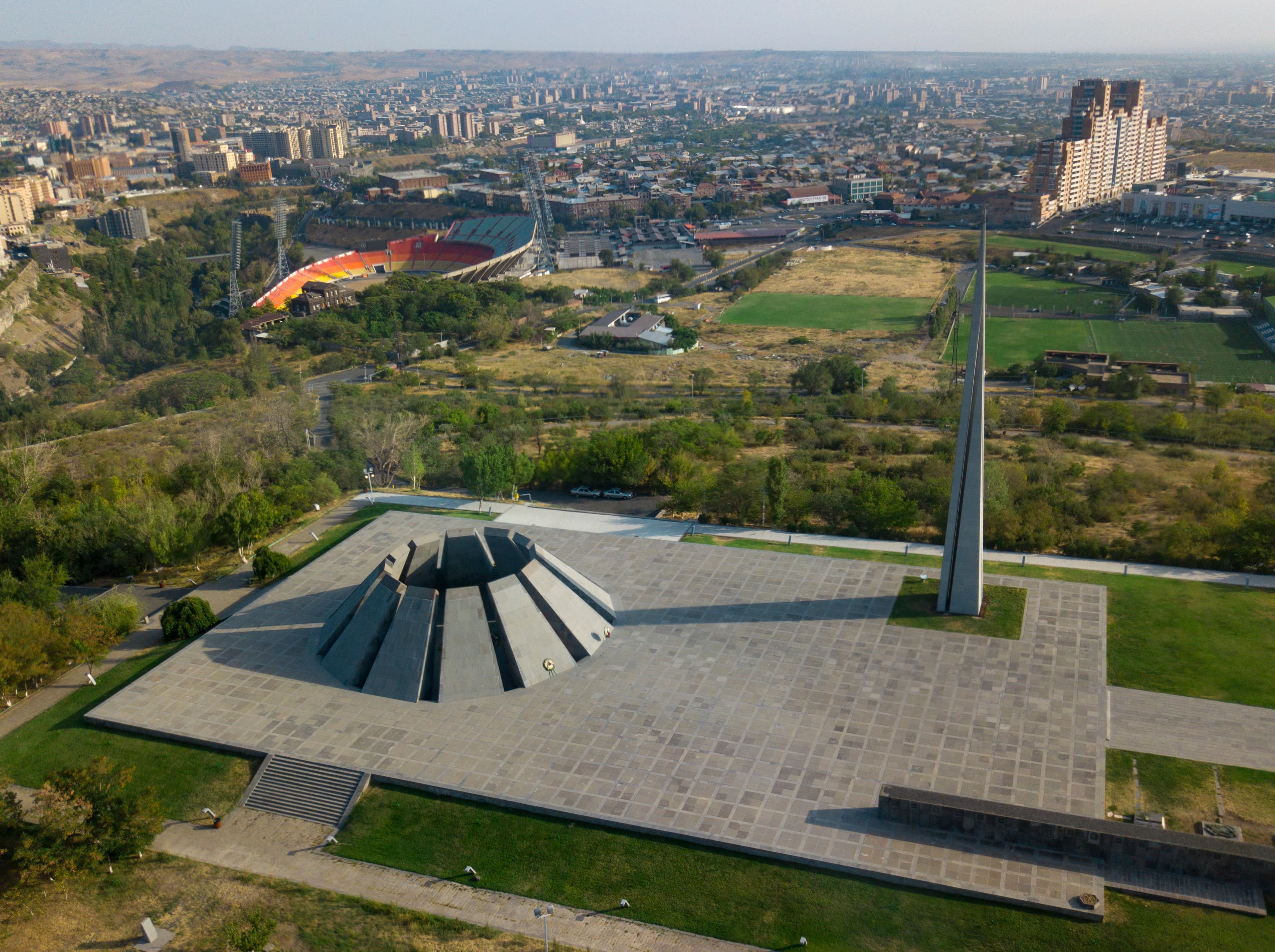 Aerial view of the Tsitsernakaberd memorial complex: the tall «Reborn Armenia» stele, the inclined khachkar-like stones around the eternal flame, and the 100-metre basalt Memorial Wall along the southern edge.
