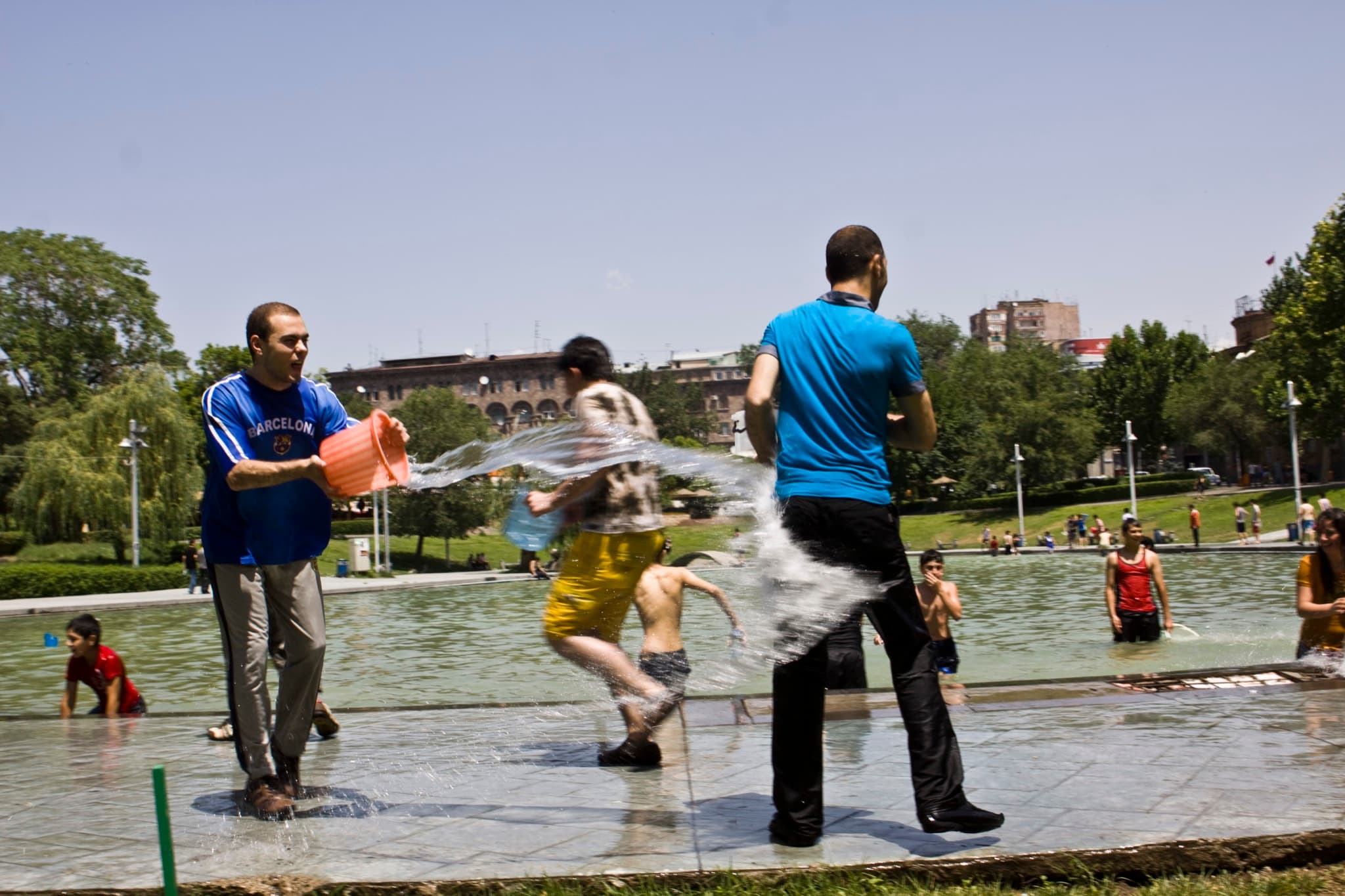 Two young men soak each other with buckets of water beside a fountain on Vardavar.